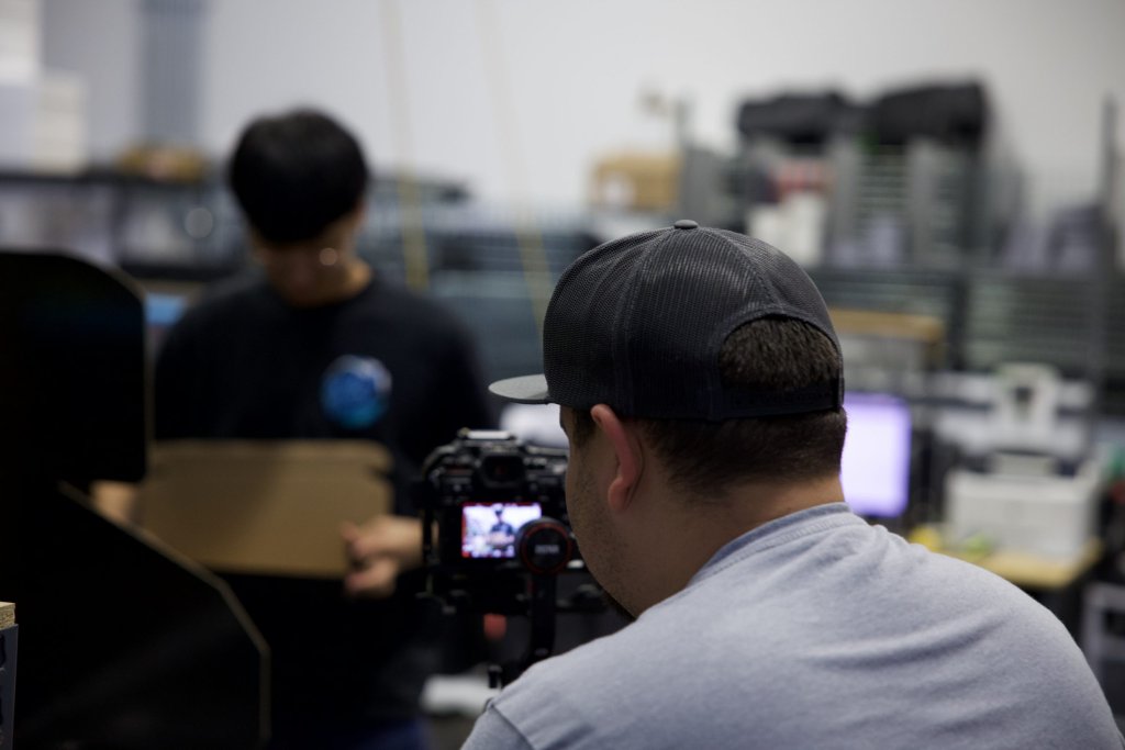 Over-the-shoulder view of a camera operator in a black cap filming a person holding a cardboard box in a workspace.