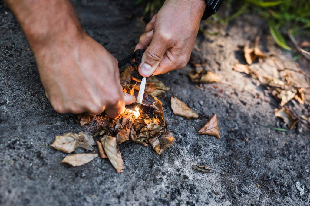 Hands striking a fire starter over a small pile of dry leaves, with sparks catching.